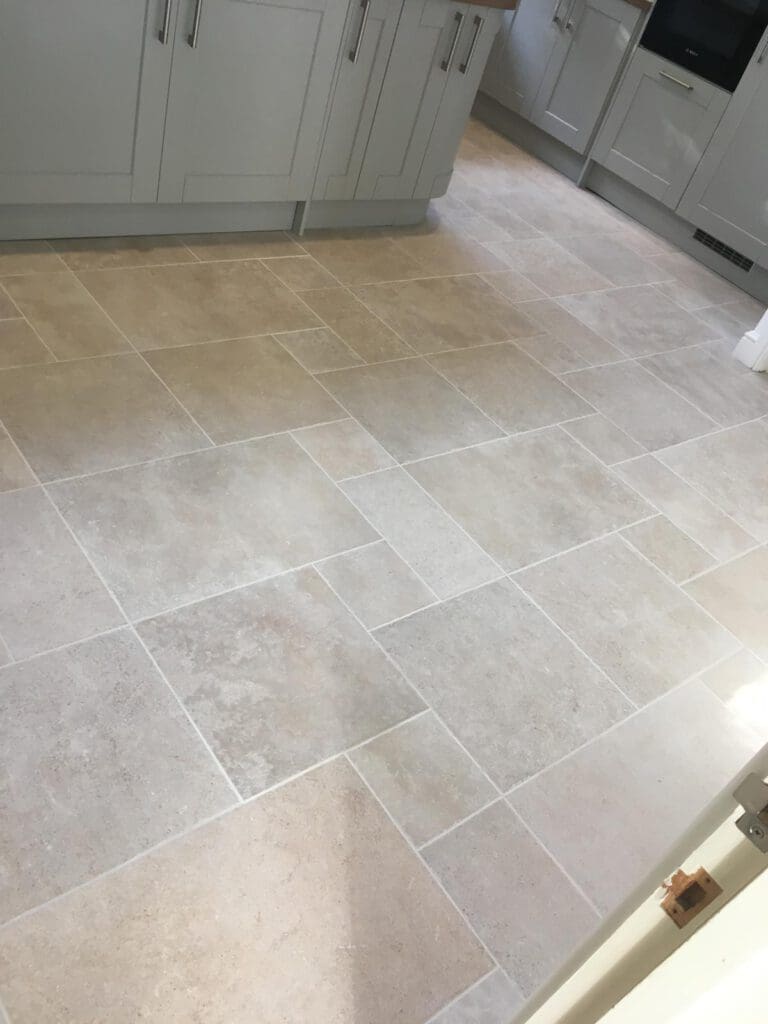 Light beige stone tiles arranged in a staggered rectangular pattern on a kitchen floor, with light grey cabinets and built-in appliances visible along the walls.