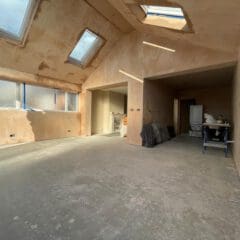 A spacious, unfinished room with plastered walls, large windows, and skylights. The floor is bare concrete, and there are some tools, furniture, and materials near the back, suggesting ongoing renovation work.