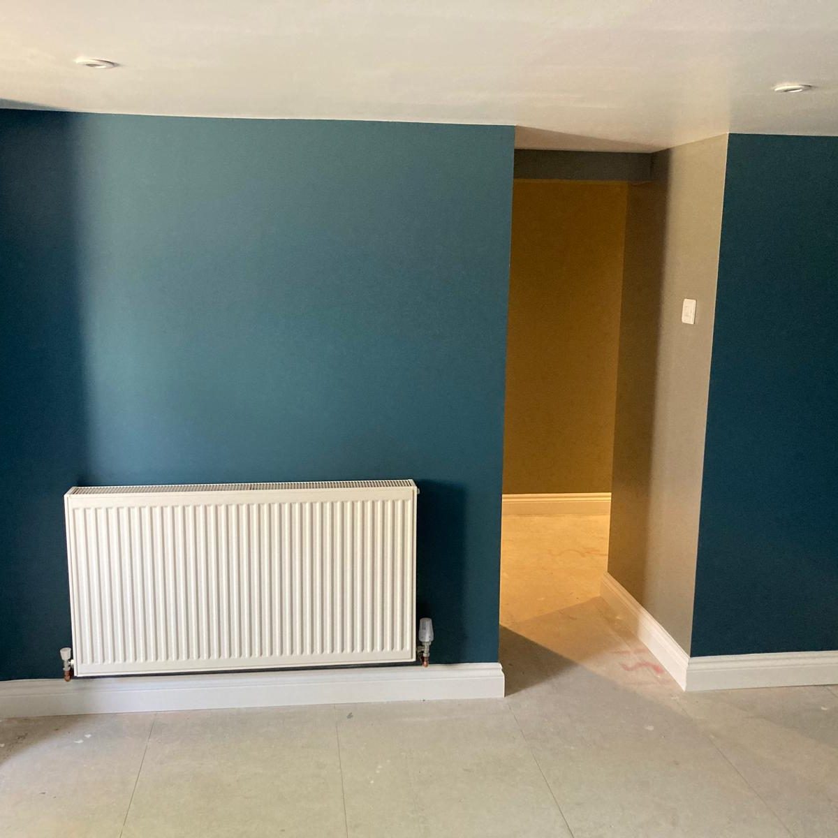 A room with teal walls, a white radiator mounted on the left wall, and an open doorway leading to a hallway with beige walls. The floor is light-colored and the ceiling has recessed lights.