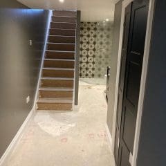 A hallway with unfinished flooring leads to a staircase with bare wooden steps. At the end of the hall, there is a wall with black star-patterned tiles and a dark door on the right. The area appears to be under renovation.