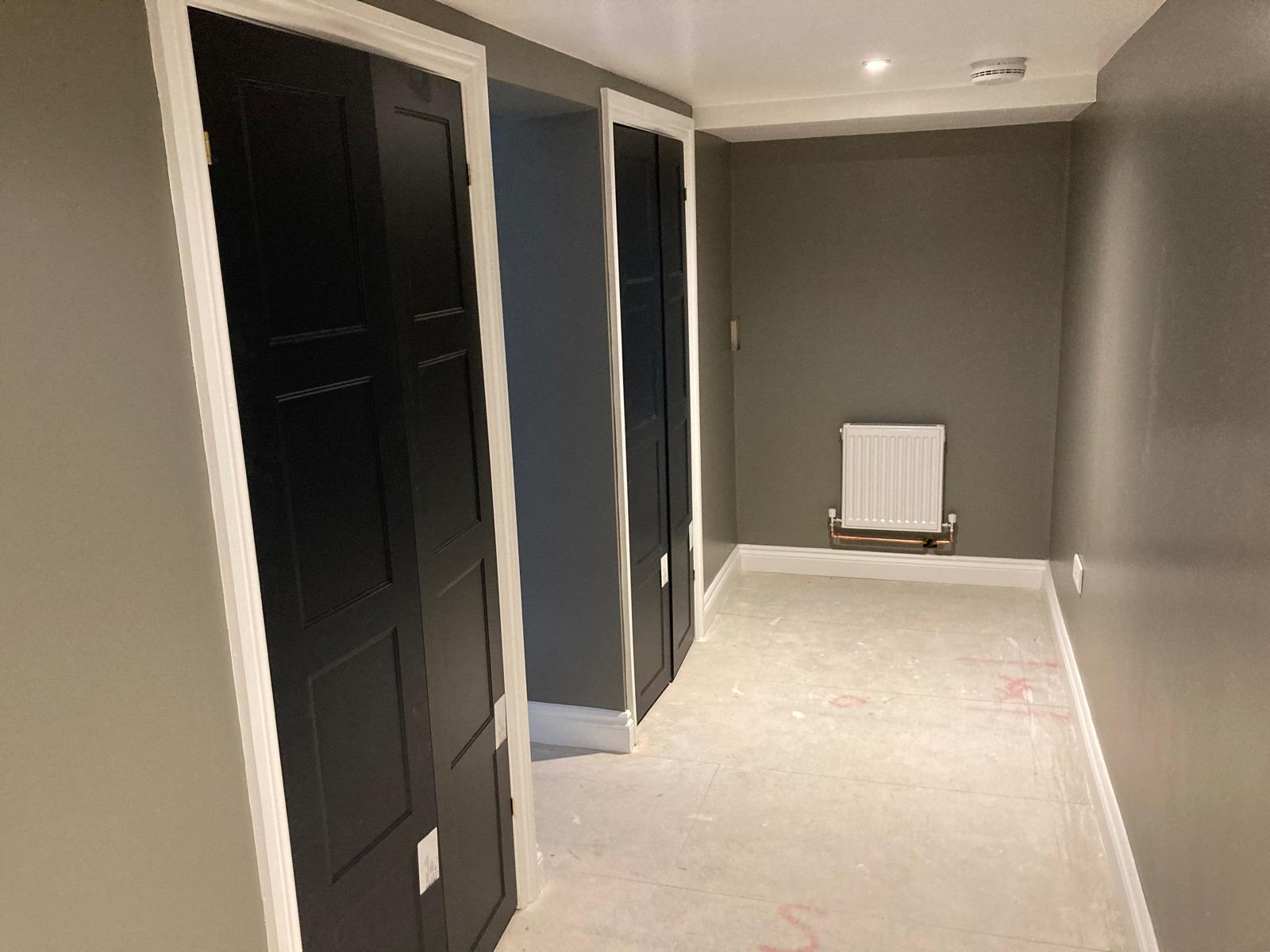 A hallway with dark paneled doors on the left, grey-green walls, light flooring, and a white radiator mounted on the wall at the end. The area appears unfinished with some markings on the floor.