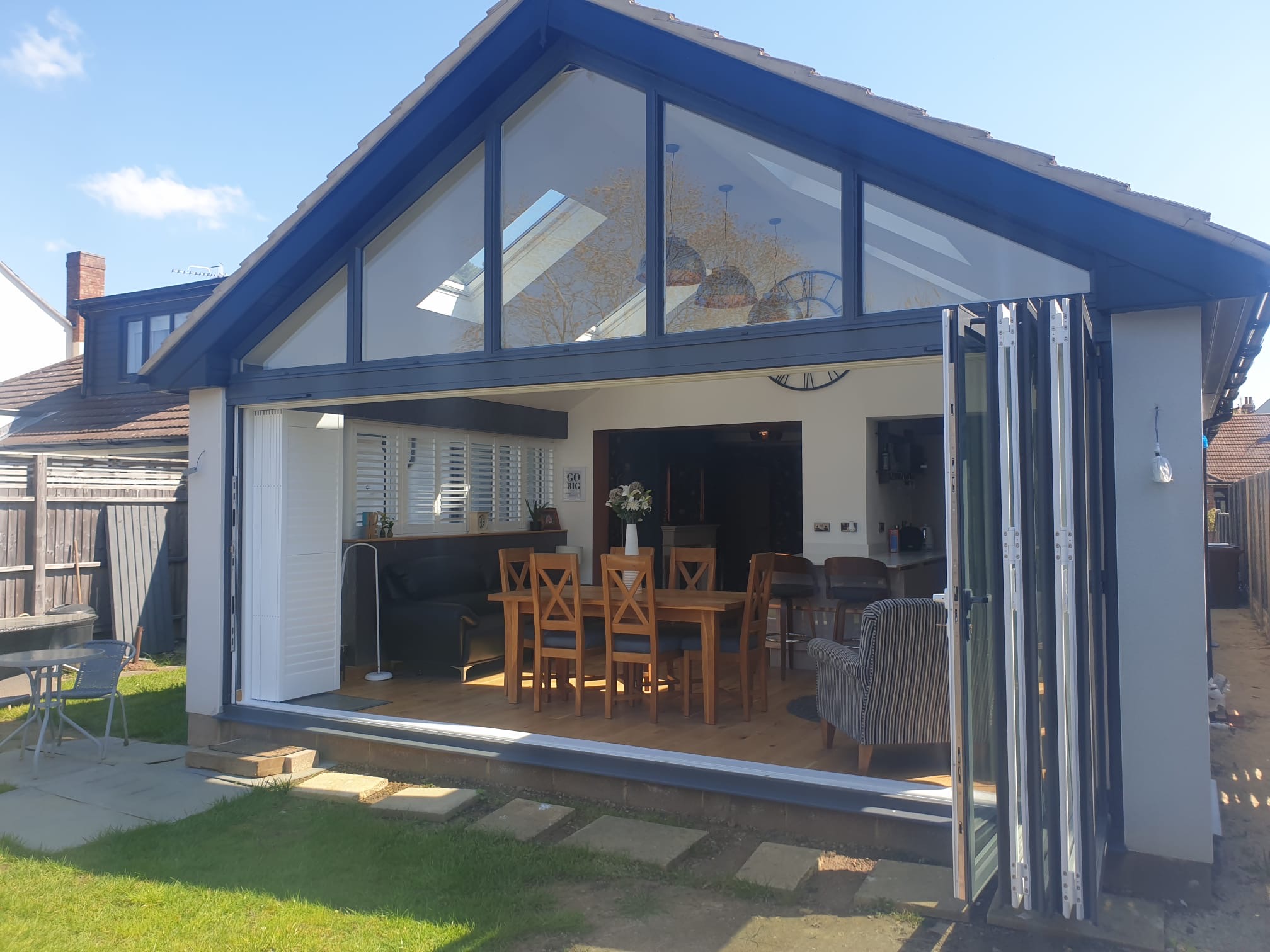 A modern house extension with large glass doors fully open, revealing a bright dining and living area with wooden furniture and skylights. The space opens onto a grassy garden patio under a blue sky.