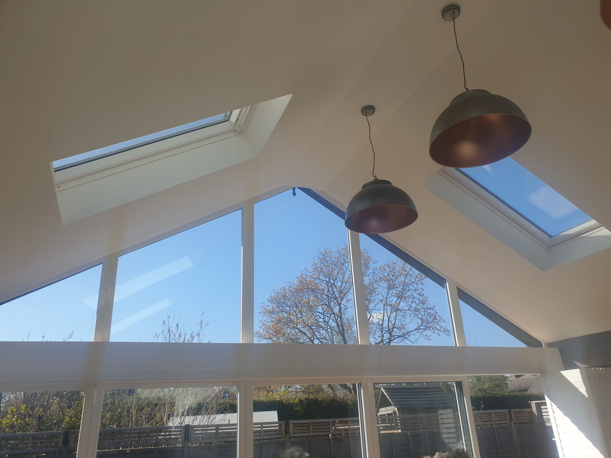 Bright, modern interior with large triangular windows, skylights, and two hanging pendant lights. Outside, trees with autumn leaves and a clear blue sky are visible, letting in lots of natural light.