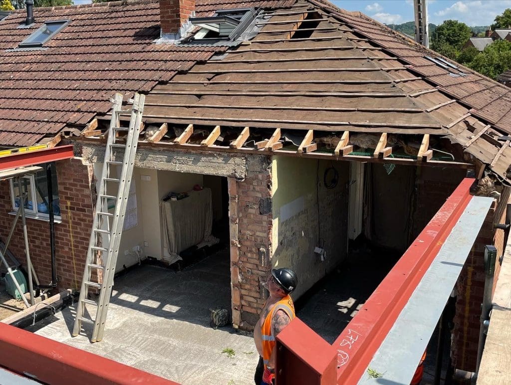 A construction worker in an orange vest and helmet stands by a partially demolished house roof, with exposed beams, bricks, and a metal frame for renovation. A ladder leans against the building on a sunny day.