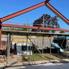 A partially constructed building with a steel frame and scaffolding stands in a backyard. Construction materials, bricks, and tools are scattered around, with trees and a clear blue sky in the background.