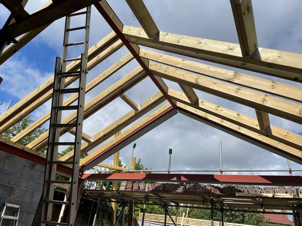 A partially constructed roof with wooden beams and a ladder leaning against the frame under a partly cloudy sky. The building's block walls are visible, and some trees can be seen in the background.