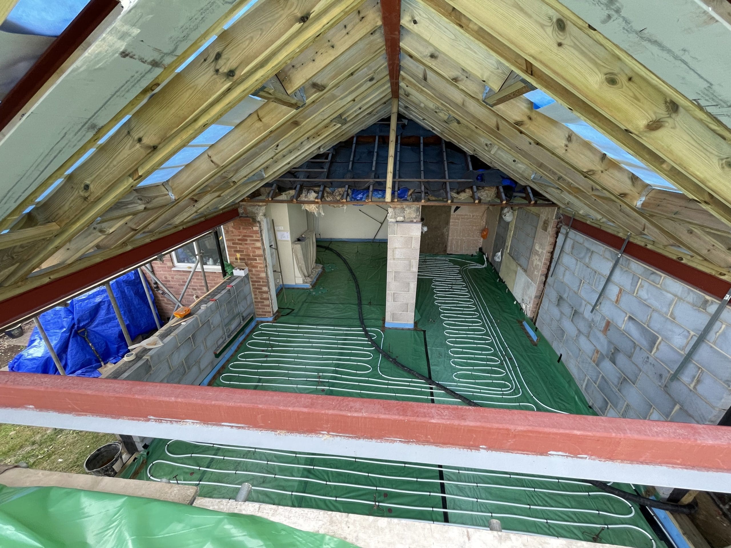 Attic view of a house under construction, showing exposed roof beams, unfinished walls, and underfloor heating pipes laid out on green insulation material. Blue tarps cover parts of the structure.