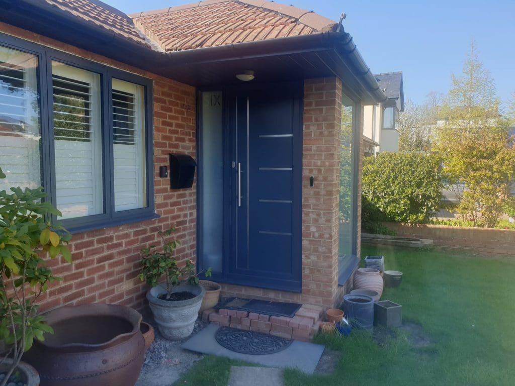 A modern house with red brick walls and a dark blue front door. Several potted plants and ceramic pots are arranged near the entrance, and a green lawn surrounds the area.